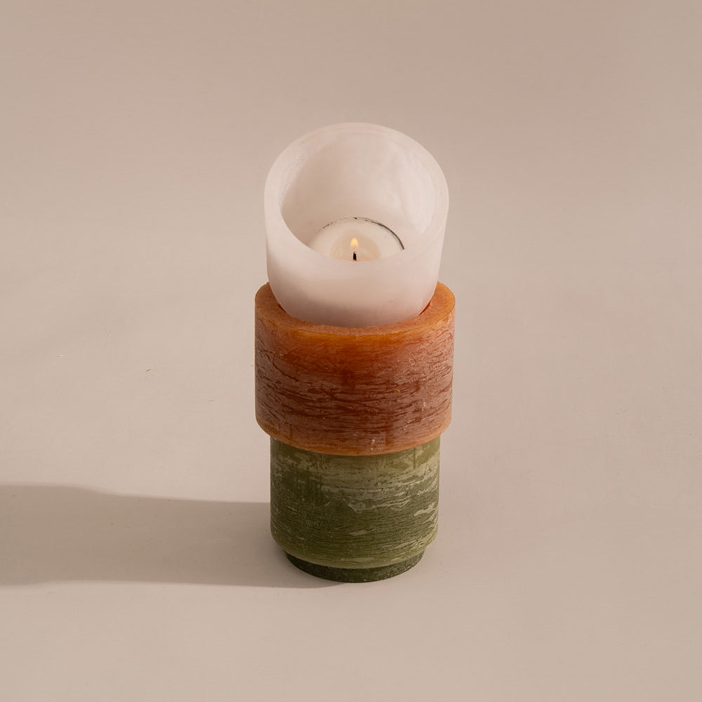 Een brandende theelichtkaars in een witte, halfdoorzichtige alabaster houder, geplaatst bovenop een gestapelde kaarsconstructie met een oranje en groene basis. De kaarsen van Stan Editions hebben een rustieke, licht verweerde textuur en werpen een zachte schaduw op de achtergrond.